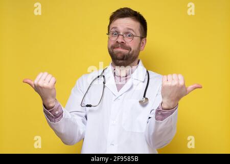 Funny doctor with a stethoscope around neck points thumbs in two directions and looks at the camera in surprise, smiling.  Stock Photo