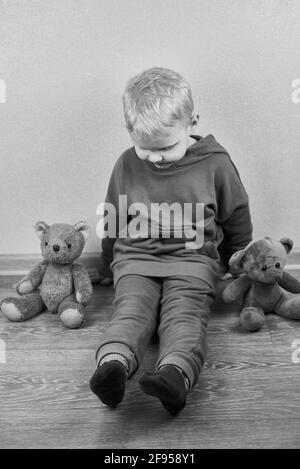 A three-year-old caucasian boy sits alone on the floor next to teddy bears, the theme of child abuse, black and white photo. Stock Photo