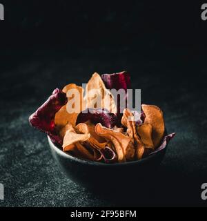 Black bowl on black background with a variation of beetroot, sweet potato and parsnip chips with copy space Stock Photo