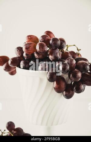 Dark red grapes in an old white porcelain bowl standing on a white table with some grapes lying on the table in the foreground. Stock Photo