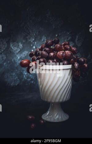 Dark red grapes in an old white porcelain bowl on dark background with some grapes lying on the table in the foreground, vertical with space Stock Photo