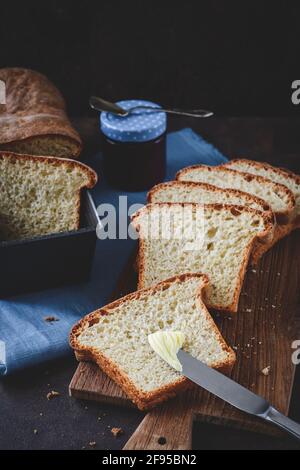 Slices of homemade white bread lying on a wooden board and blue napkin, the front spread with butter, the chunk of bread and the glass with jam in the Stock Photo