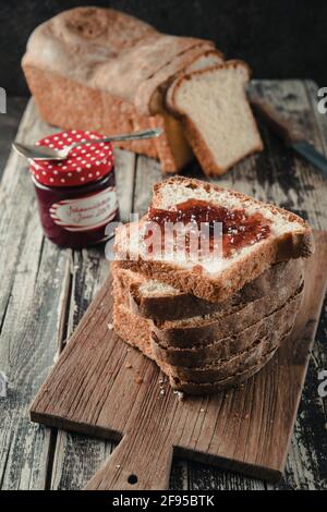 Stacked slices of homemade white bread lying on a wooden board, the uppest spread with jam, the chunk of bread and the glass with jam in the backgroun Stock Photo