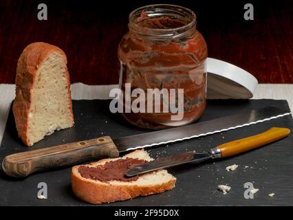 A slice of white bread, cut from a larger loaf, is spread with gianduia cream on a dark background Stock Photo
