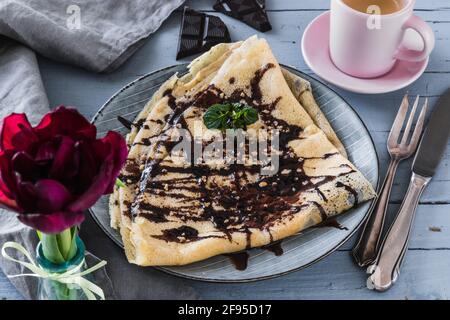 French pancake with chocolate, brittle and mint, decorated with espresso in a rose cup, dark red tulip, grey napkin and cutlery lying on a light grey Stock Photo