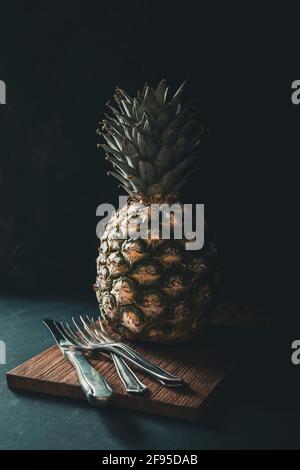 Central whole pineapple on wooden board with a knife and three forks on a dark background. Vertical. Stock Photo