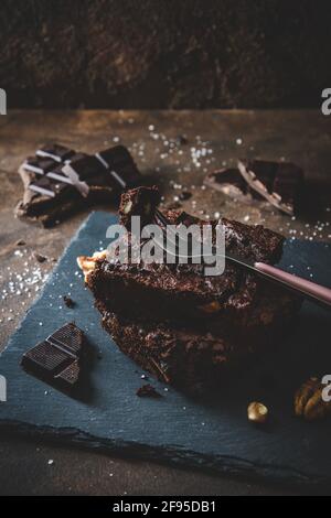 Two pieces of walnut brownies, pieces of dark chocolate, salt and fork on slate platter and dark brown background Stock Photo