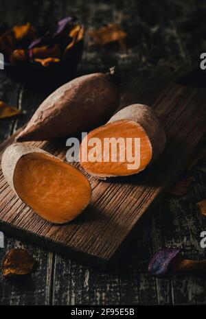 One whole, one devided sweet potatoes lying on a dark wooden board, vertical with vegetables chips in the dark background. Stock Photo