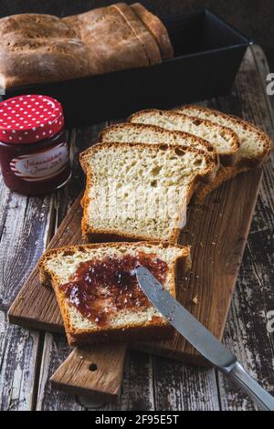 Slices of homemade white bread lying on a wooden board on a wooden table, the front spread with jam, the box with bread and the glass with jam in the Stock Photo