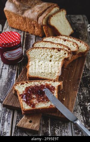 Slices of homemade white bread lying on a wooden board, the uppest spread with jam, the chunk of bread and the glass with jam in the background. Verti Stock Photo