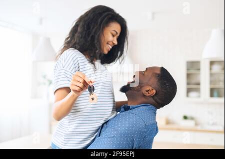 African woman holding home keys while hugging boyfriend in new apartment, lovely couple feels ecstatic about their recent purchase of new home which makes them feel being on the verge of happiness Stock Photo