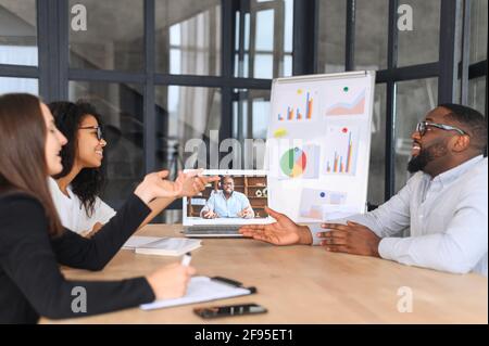 Lively team holds meeting with black boss via video link in modern office, teleconference meeting using laptop, african businessman having video call with his business team online, talk about strategy Stock Photo