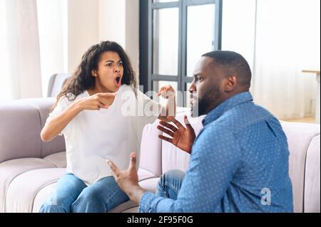 Stressed young married family couple arguing emotionally, blaming lecturing each other, sitting on couch. Depressed husband quarreling with wife, having serious relations communication problems. Stock Photo