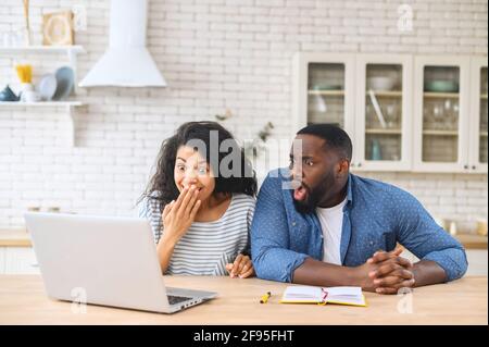 Overjoyed young multiracial family couple looking at the laptop monitor, feeling excited of getting online lottery win notification, reading unexpected news, feeling surprised together at home Stock Photo