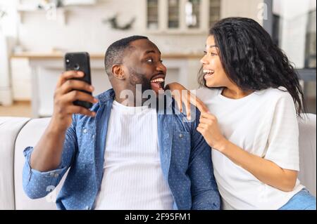Smiling millennial black couple sit on couch in kitchen have fun browsing Internet on modern smartphone together, happy young husband and wife relax on sofa use cellphone enjoy family weekend at home Stock Photo