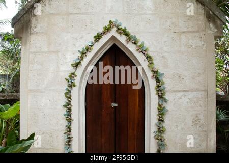 A slightly ajar old-fashioned medieval frame door in Barbados, surrounded by climbing green ivy vines with blossoms and old-fashioned colonial chapel. Stock Photo