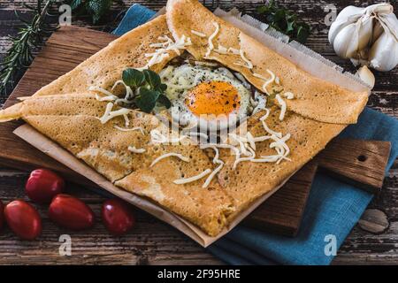 French pancake crepe with fried egg, herbes, tomatoes and cheese on a slat on blue linen on a wooden table. Closeup. Stock Photo