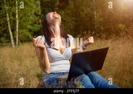 happy young woman with laptop on a sunny lawn. freelancer works in nature. student studies remotely on nature landscape outdoor Stock Photo