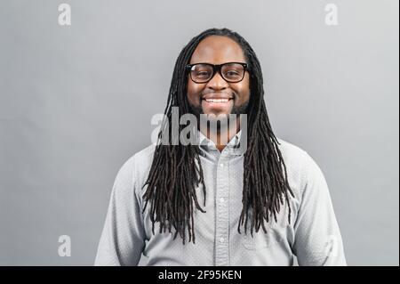 Smiling young handsome African American guy with dreadlocks in smart casual shirt and eyeglasses standing isolated over grey background and looking at the camera, cheerful optimistic black man Stock Photo