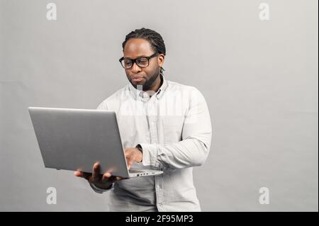 Smart and concentrated African-American businessman wearing smart casual shirt and eyeglasses using a laptop, a male office employee holding computer and typing message, isolated on grey Stock Photo