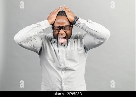 Mad frustrated black guy holding head and screams isolated on grey, angry African-American man with suffering face expression feels disappointment and depression, yelling with eyes closed Stock Photo