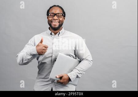 Cheerful African-American man carrying laptop computer isolated on grey. Happy black male entrepreneur with laptop showing thumbs up, recommending app for small business Stock Photo