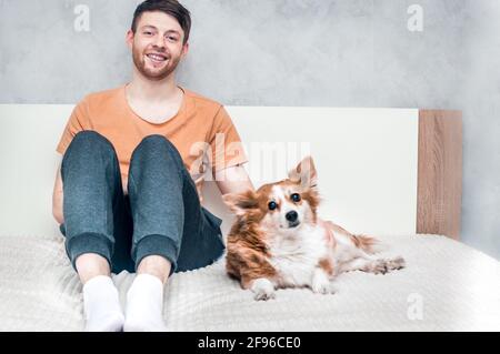 young guy is sitting on the bed with his ginger dog. Close-up portrait. Owner and dog Stock Photo