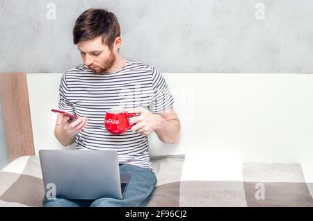 young man works on a laptop with a phone in his hand and drinks coffee from a red big cup on the bed at home.Concept working remotely Stock Photo