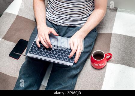 Young man working on a laptop in bed at home and big red cup beside.A man looks at a smartphone. Concept Working remotely Stock Photo