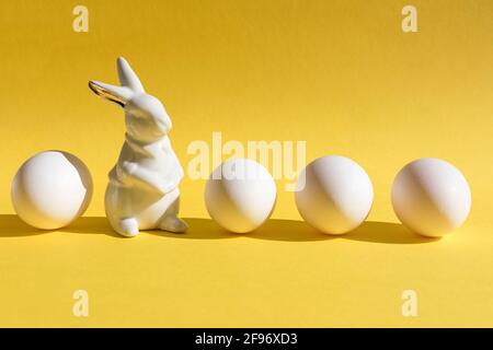 Creative minimalist Easter concept. Porcelain rabbit with four eggs on yellow background. Trendy colors of 2021. Copy space. Stock Photo