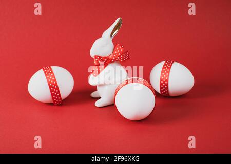 Easter Concept. Porcelain rabbit with three white eggs decorated with red ribbon on red paper background. Copy space. Stock Photo