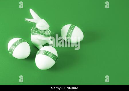 Easter Concept. Porcelain rabbit with three white eggs decorated with green ribbon on green paper background. Copy space. Stock Photo