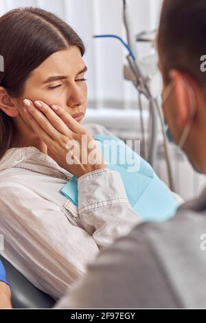Young woman having toothache, sitting in a dental chair at the clinic. Female patient with terrible toothache visiting dentist. Stock Photo