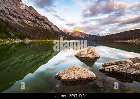 A view looking south across Medicine Lake n Jasper National Park with boulder in the water in the foreground and partially cloudy skies above Stock Photo