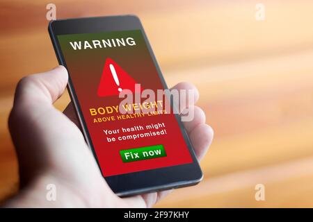 Closeup of male hand holding modern mobile phone with red screen and warning symbol with text warning about overweight. Weight loss, concept of obesit Stock Photo