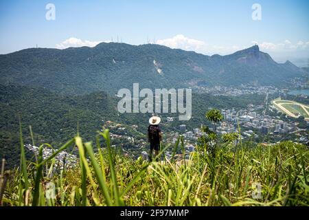 A local man with a hat standing at the edge of the Two Brothers Hill (Morro Dois Irmãos) in Rio de Janeiro City - enjoying the view to the Corcovado with its Statue of Jesus atop its peak, entitled Christ the Redeemeer and the Tijuca Forest National Park. Shot with . Stock Photo