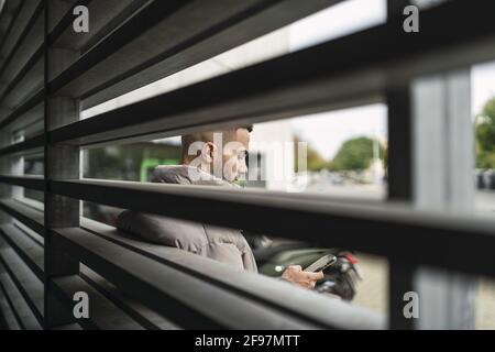 Shallow focus shot of a young European man scrolling on his phone behind a fence Stock Photo