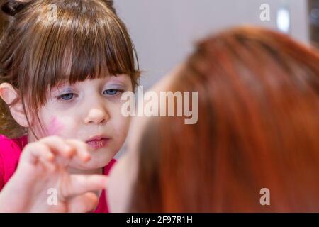 Portrait of a cute, blue-eyed, brown-haired girl with makeup in her face playing to put makeup in a redhead woman Stock Photo