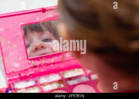 A cute blue-eyed girl with makeup on her face smiling and looking at a mirror in a makeup set Stock Photo