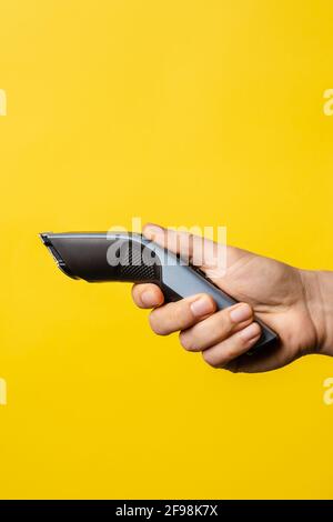 Close up on hand of unknown caucasian man holding electric beard trimmer cordless hair cutter in front of modern yellow background Stock Photo