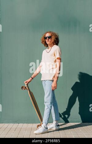 vertical portrait of a caucasian young skateboarder looking at camera. He is wearing casual clothes and sunglasses. Green wall background Stock Photo