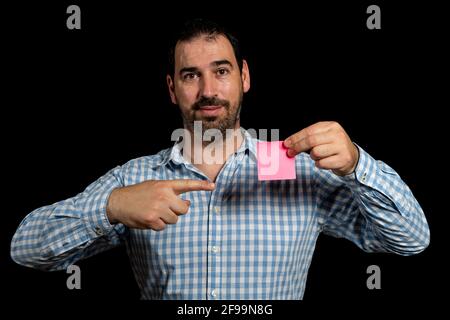 Handsome bearded man in a white and blue plaid shirt pointing a posit with one hand and holding ir with the other while smiling looking at the camera Stock Photo