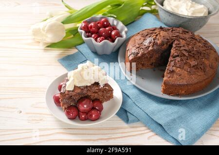 Homemade chocolate cake and bowls with morello cherries and whipped cream, some tulip flowers and a blue napkin on a bright wooden table, copy space, Stock Photo