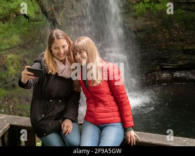 Two young women take selfies in front of a waterfall in Ireland Stock Photo
