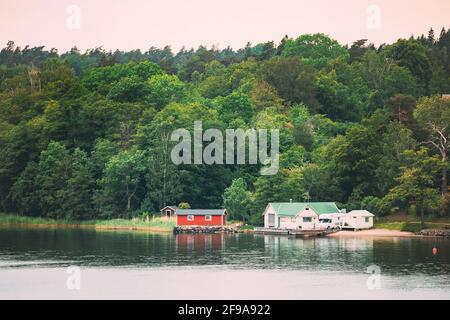 Sweden. Beautiful Red Swedish Wooden Log Cabin House On Rocky Island Coast In Summer. Lake Or River And Forest Landscape Stock Photo