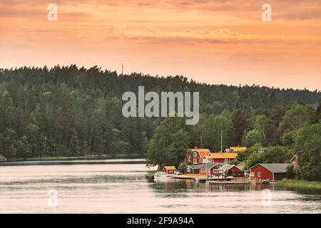 Sweden. Many Beautiful Red Swedish Wooden Log Cabins Houses On Rocky Island Coast. Lake Or River Landscape Stock Photo