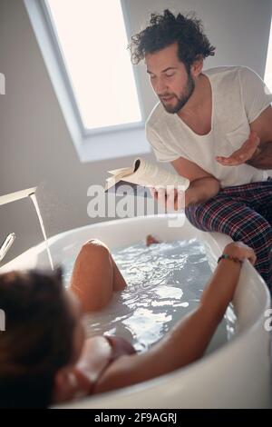 romantic moment of lovely caucasian couple in the bathroom. beardy gut is reading a book to his shy partner in the bathtub, sharing a moment. Valentin Stock Photo