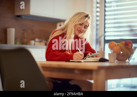 beautiful young adult caucasian blonde female writing down plans for the day, casually dressed, sitting in sunny kitchen
