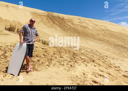 A tour guide stands in front of the giant sand dune at Te Paki, Northland, New Zealand. He holds a sandboard which can be used to ride down the dune Stock Photo