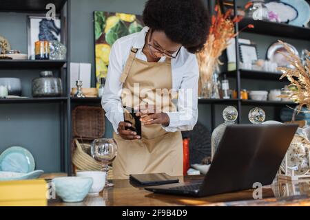 African saleswoman in eyeglasses taking picture of all assortment of decor shop on modern smartphone. Pretty woman in apron creating online catalogue using wireless laptop. Stock Photo